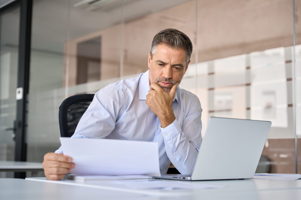 Doubtful serious busy mid aged mature professional business man ceo manager executive holding corporate financial documents reading paper letter sitting at desk in office feeling doubt uncertainty.