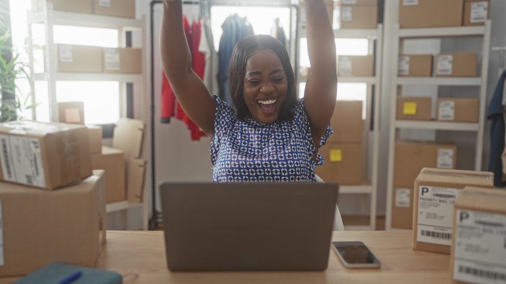 Woman at laptop raising fists in celebration among cardboard shipping boxes, smartphone and shelving in a building packing area; small business success joy.